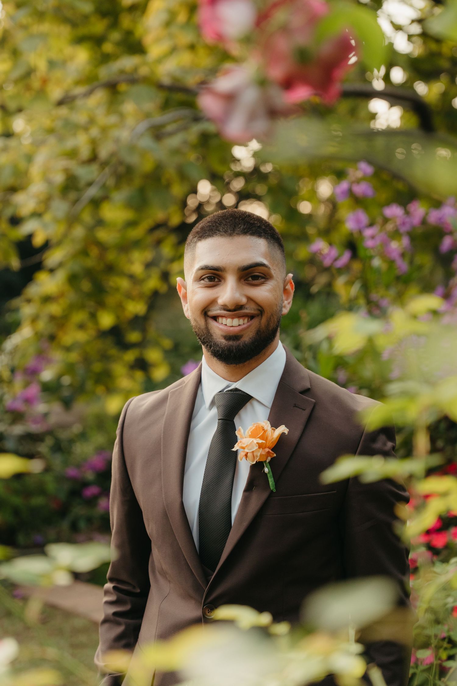 Smiling groom in brown suit with orange boutonniere poses in lush garden with colorful flowers.
