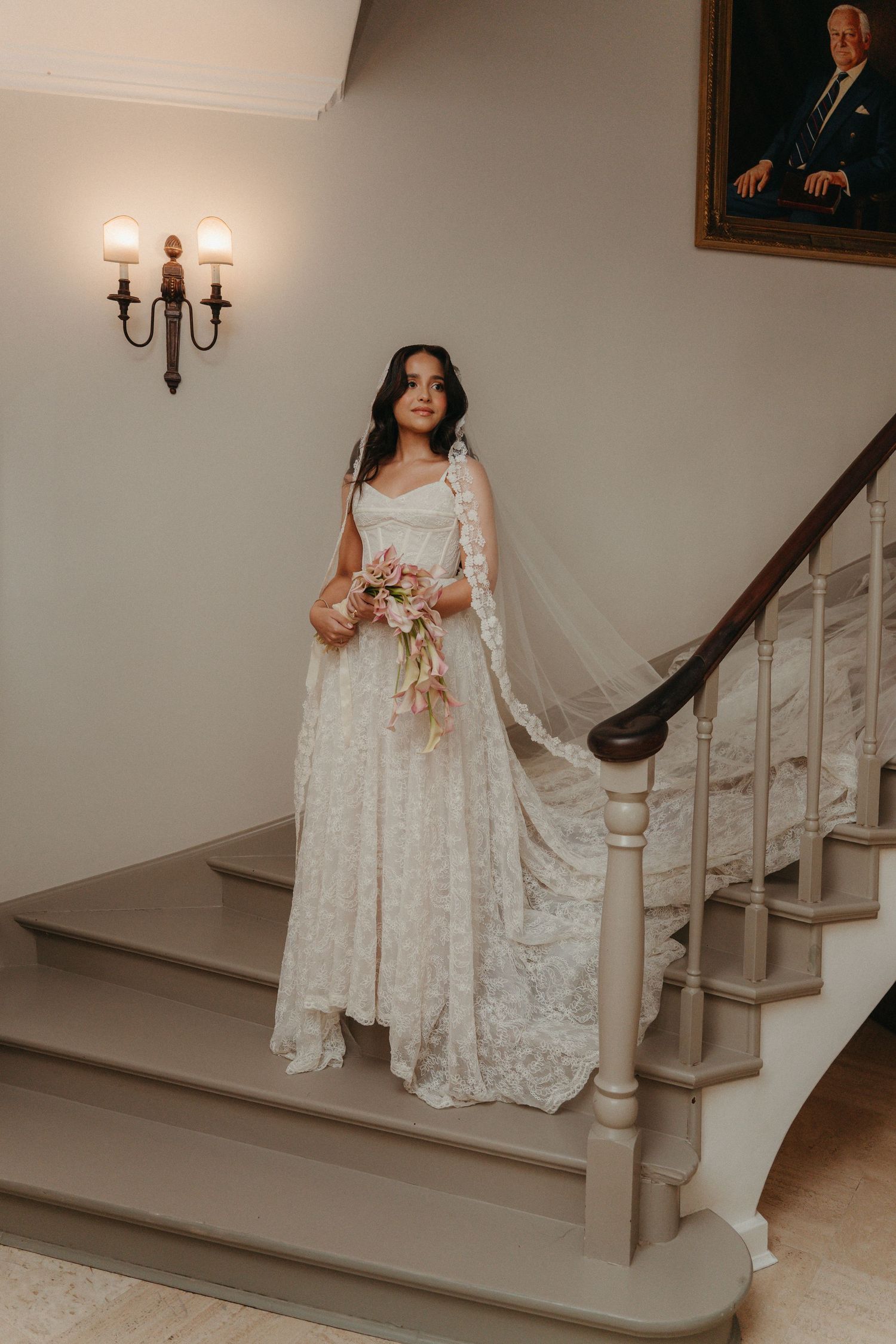 Bride in lace wedding gown holding bouquet on elegant staircase with wall sconce and portrait in background.