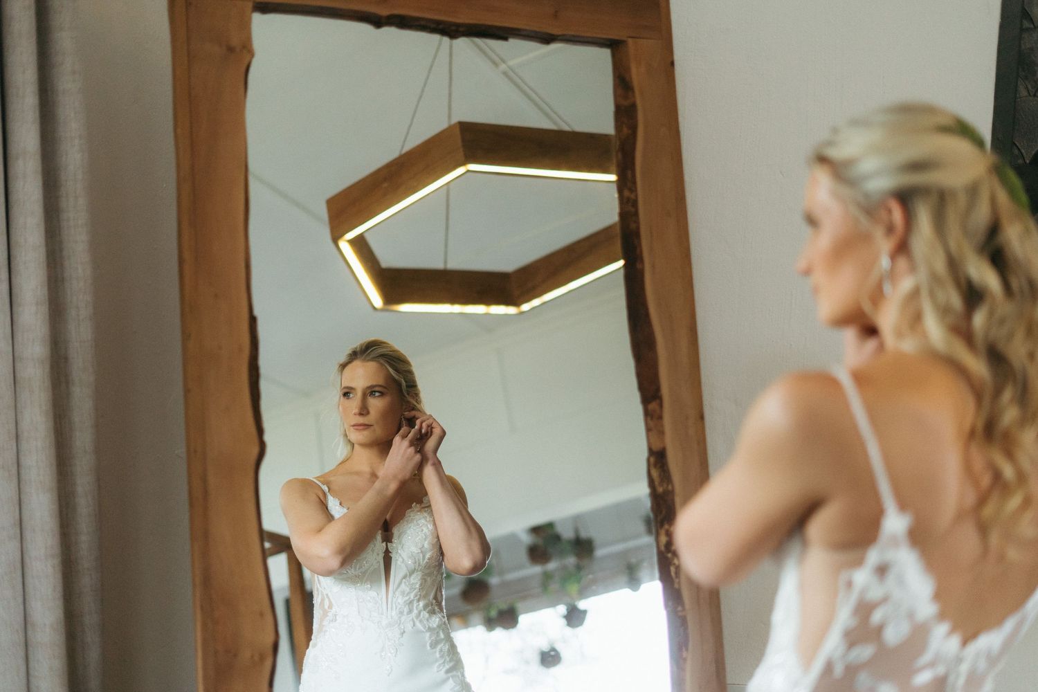 Bride adjusting earring while getting ready, reflected in a rustic wooden mirror with hexagonal light fixture.