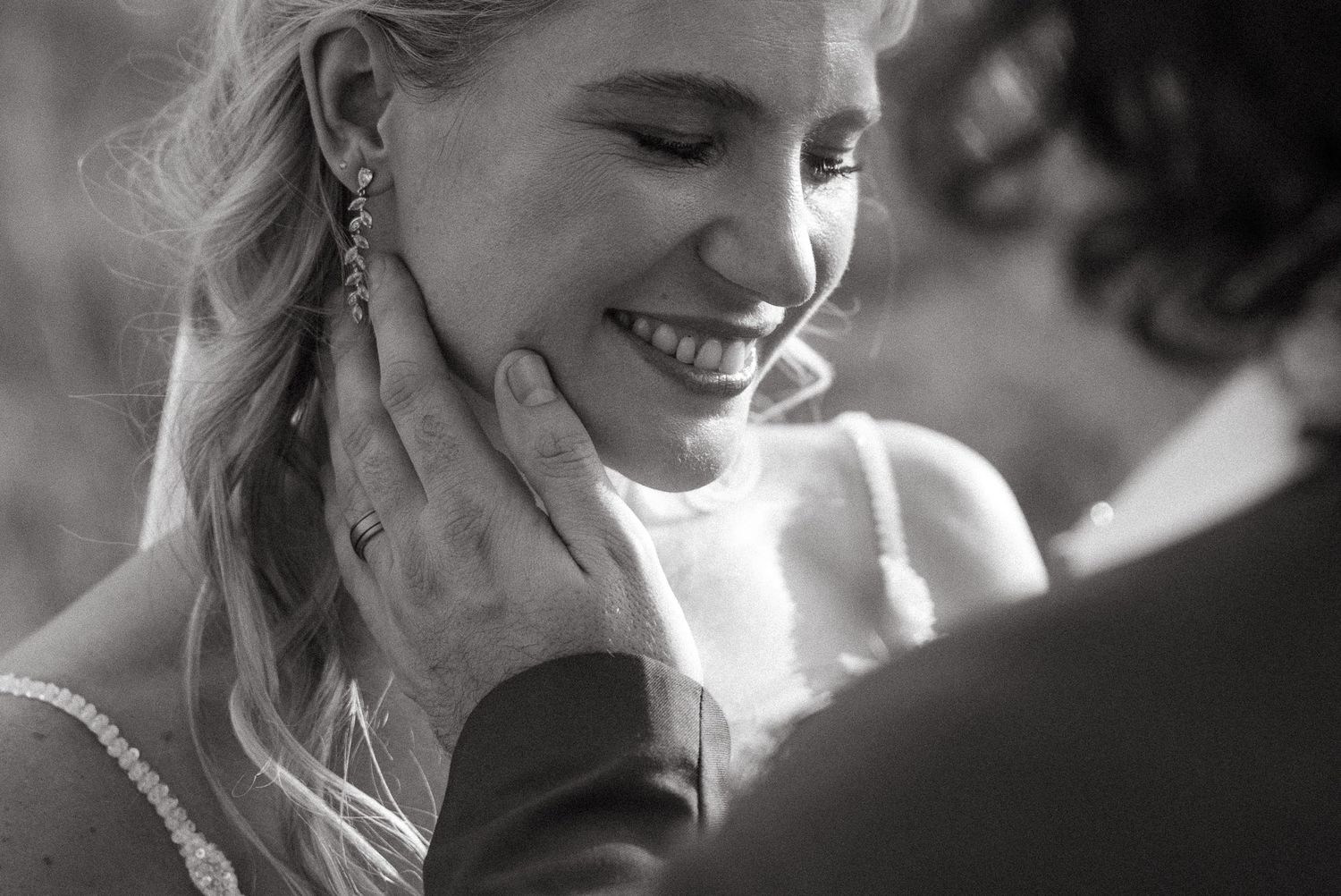 Black and white wedding photo of a smiling bride as groom tenderly cups her face, showcasing elegant earrings and ring.