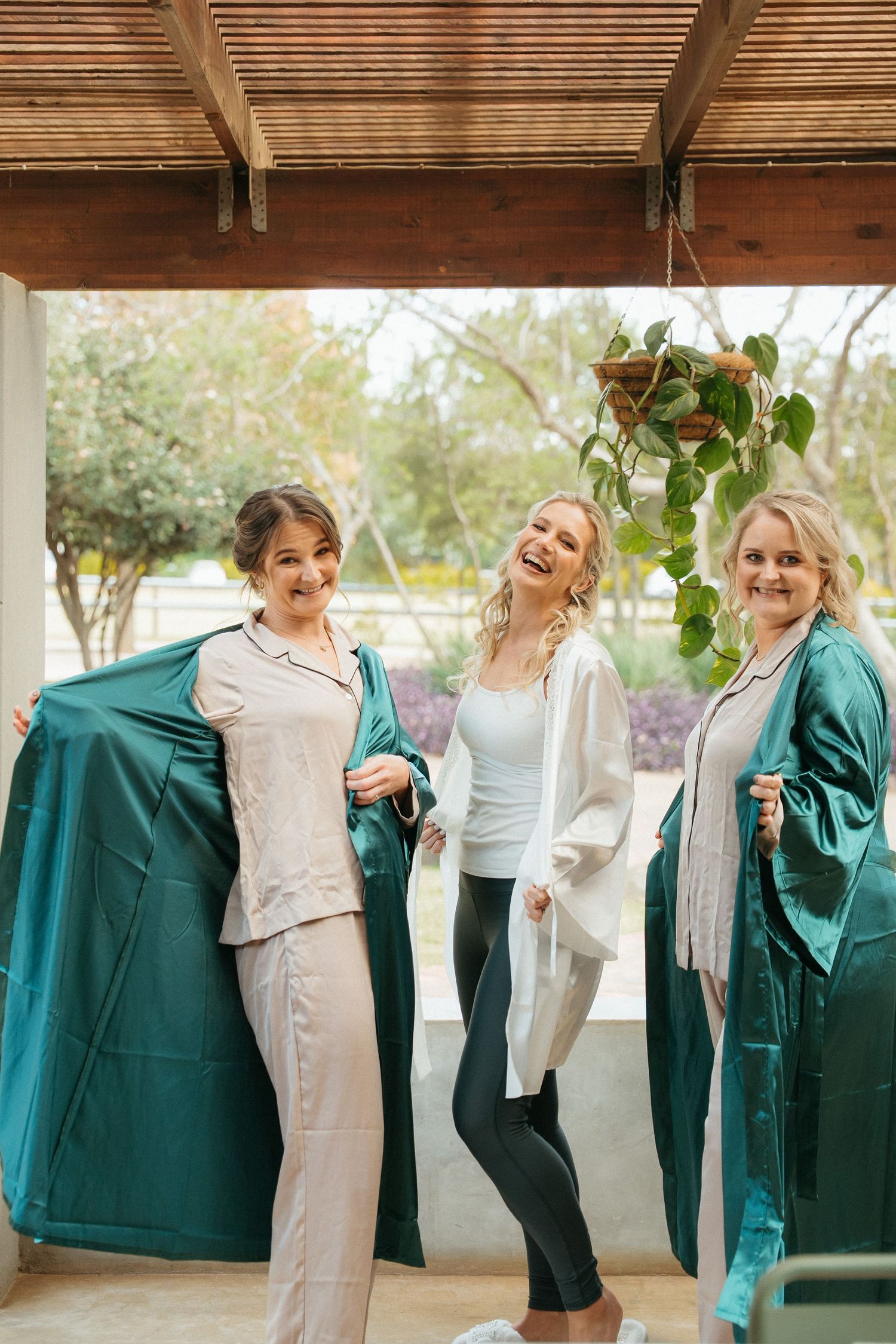 Three women laughing in teal satin robes and pajamas on a covered patio, enjoying a fun bridal party morning.