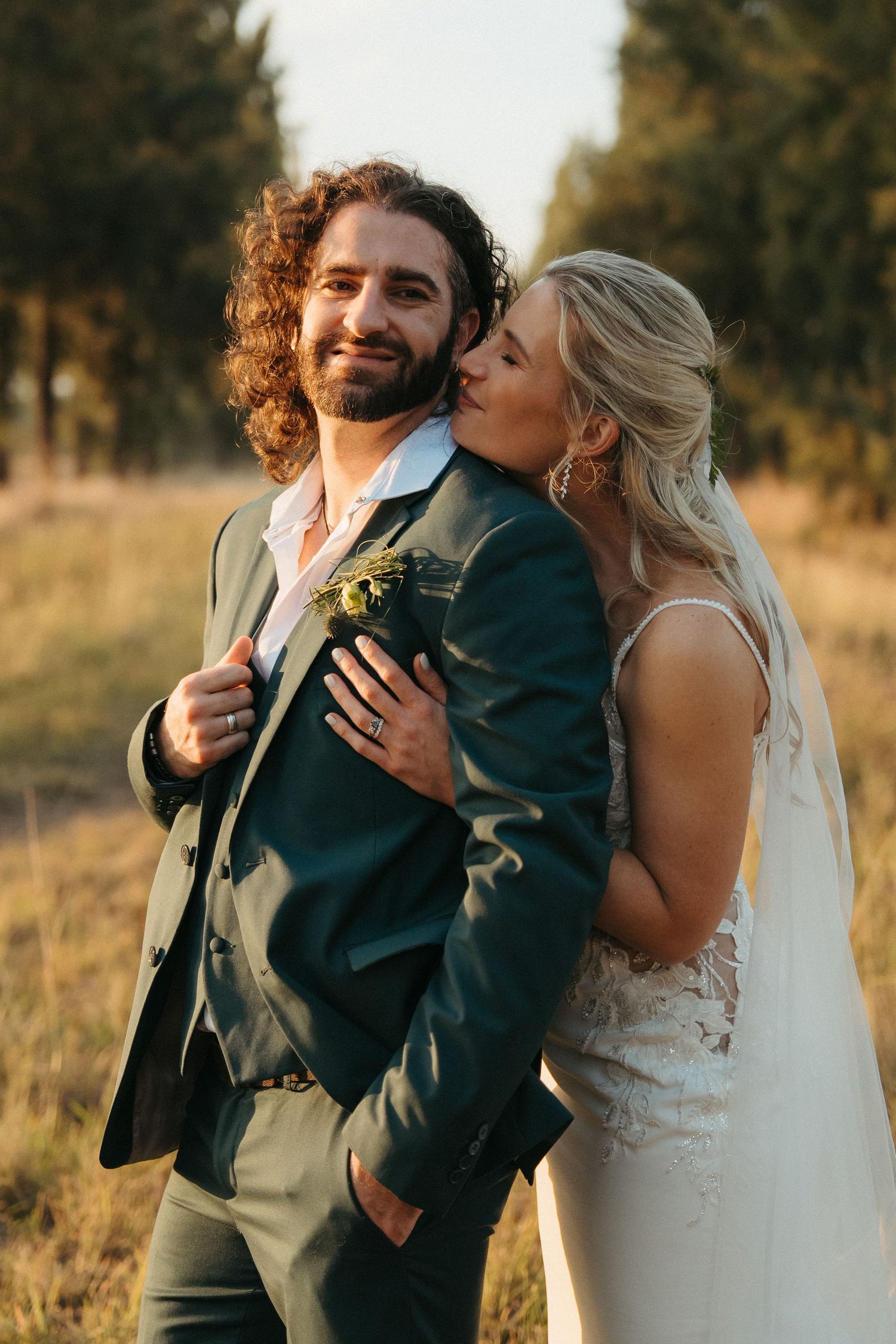 Bride in lace gown kissing groom in green suit on cheek during outdoor golden hour wedding portrait.