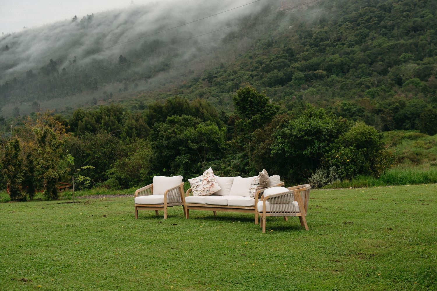 Outdoor wooden sofa set with cream cushions on lush green lawn against misty mountain forest backdrop in Plettenberg Bay.