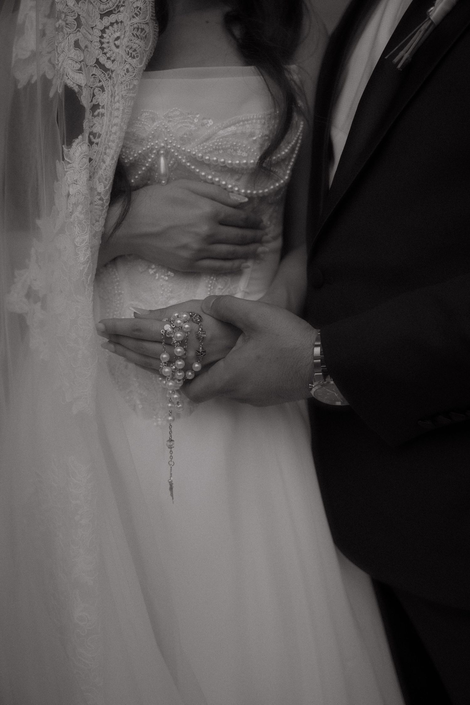 Black and white wedding photo of bride and groom holding rosary beads, bride in lace gown with veil.