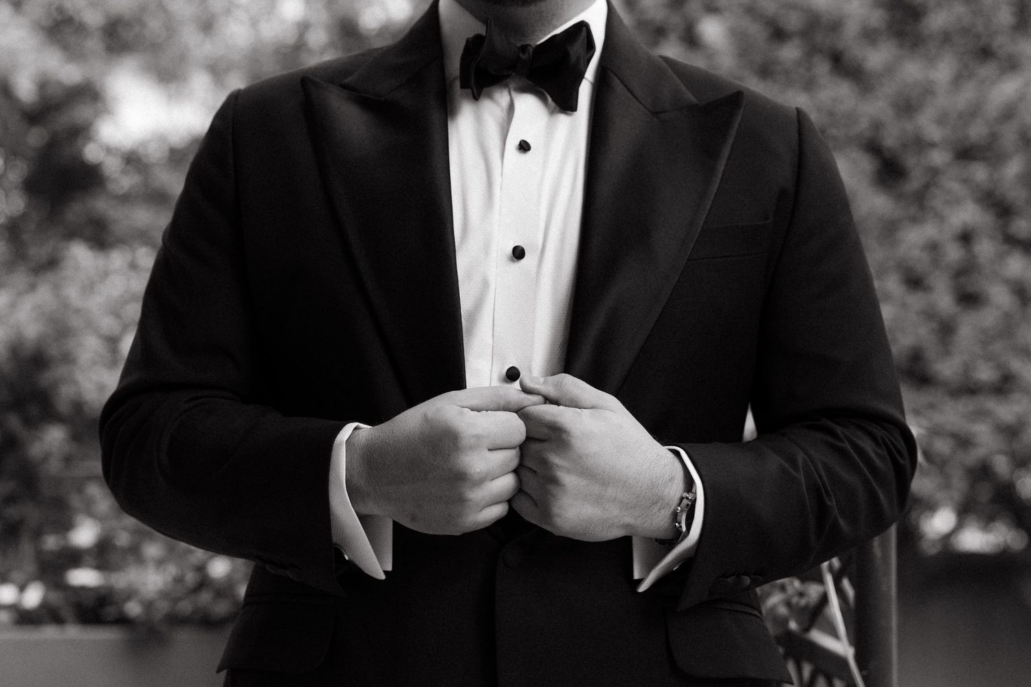 A man in a black tuxedo with bow tie adjusts his jacket buttons in a classic black and white portrait photo.