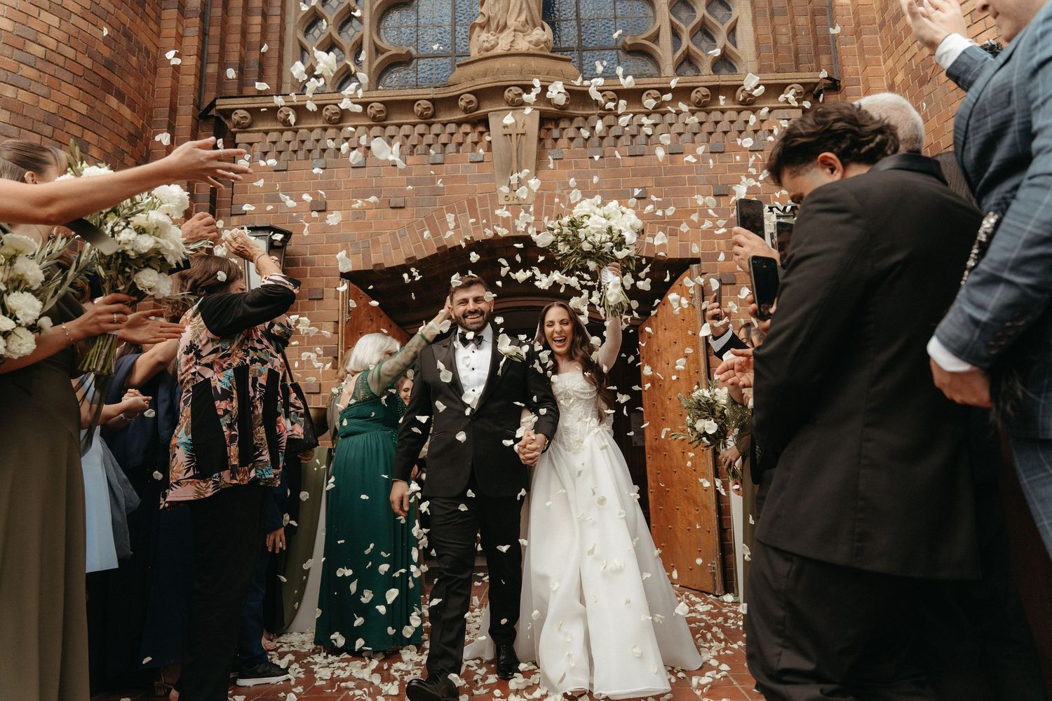 Bride and groom walk through confetti shower outside Rosebank Catholic Church, Johannesburg surrounded by cheering wedding guests.