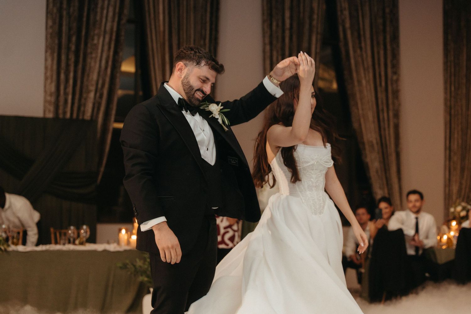 Bride and groom share first dance at elegant wedding reception, groom spinning bride in white gown under warm candlelight.