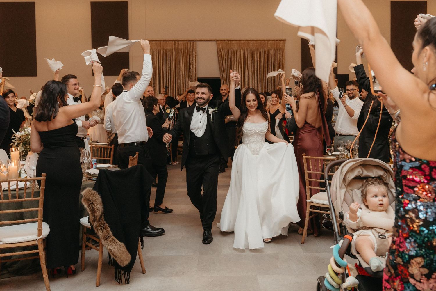 Bride and groom walk triumphantly through cheering wedding guests waving white handkerchiefs in elegant reception hall.