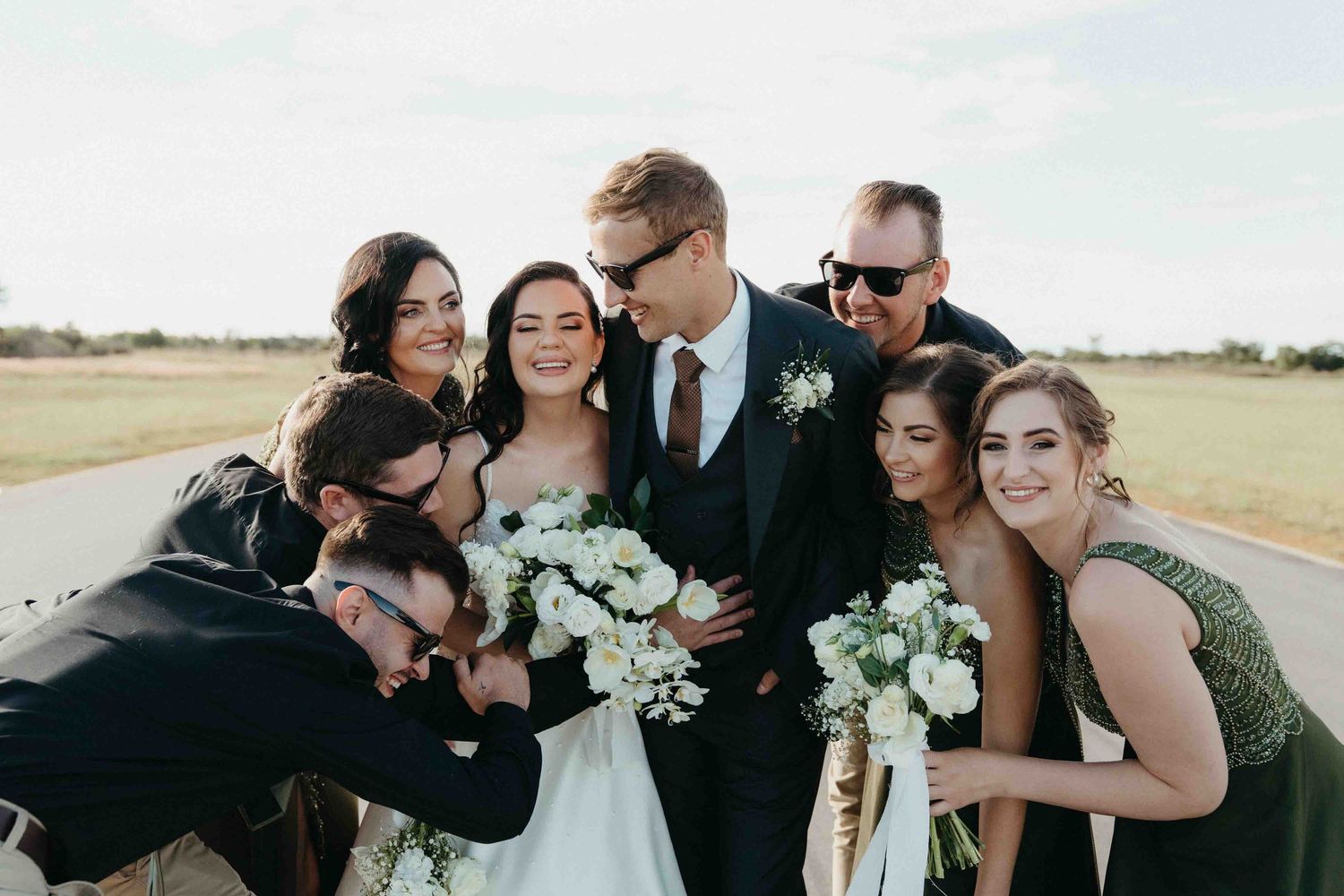 Wedding couple kissing surrounded by smiling bridesmaids and groomsmen outdoors on a sunny day.