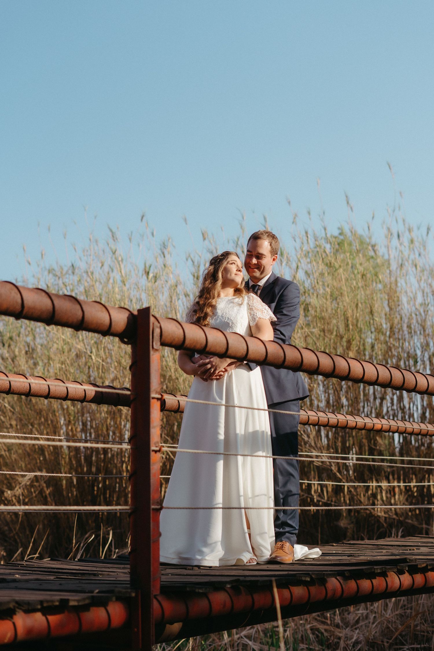 A romantic moment shared between a couple on a rustic bridge surrounded by tall reeds and blue skies.