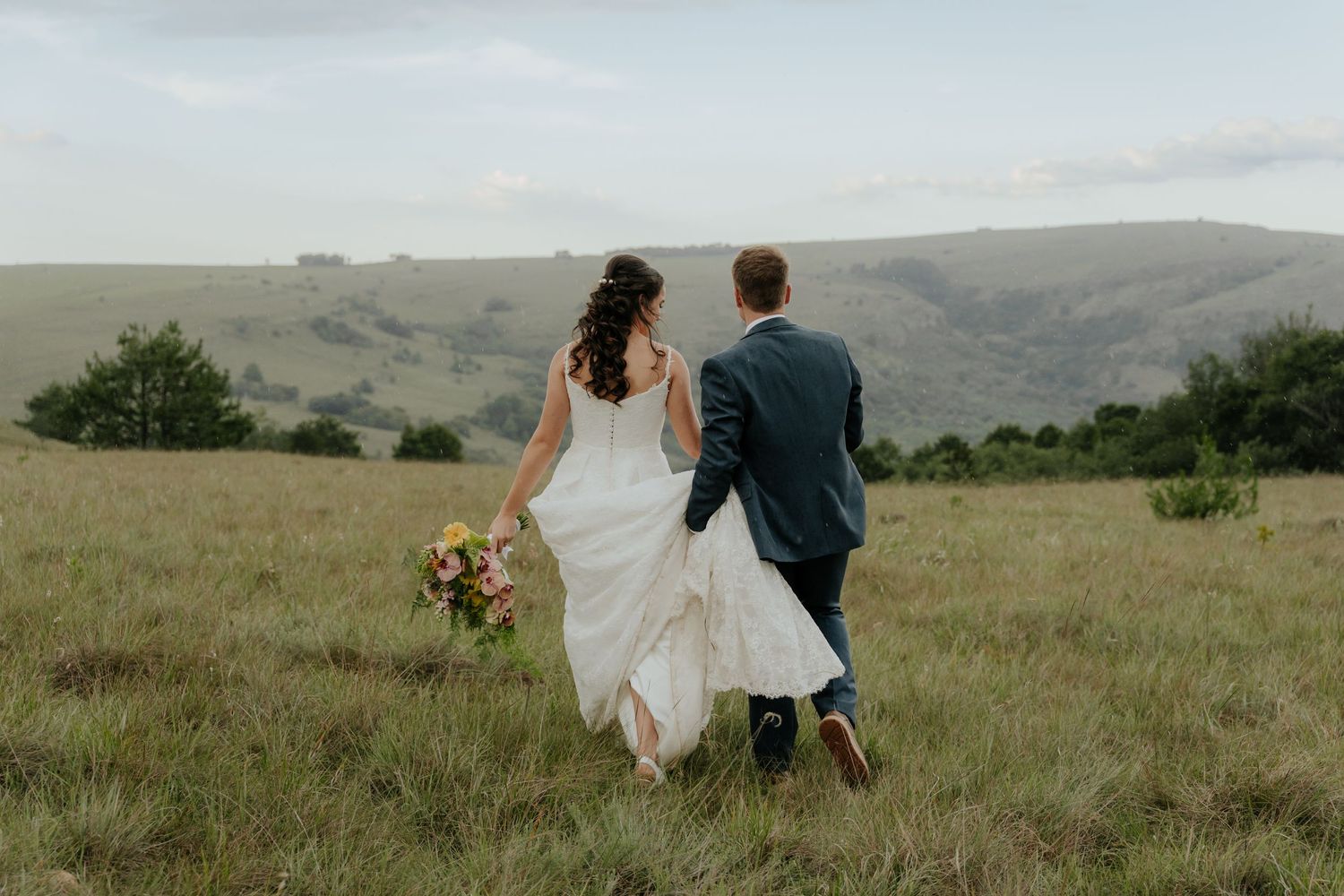 Bride and groom walking hand in hand through lush green meadow with stunning Mpumalanga mountain landscape behind.