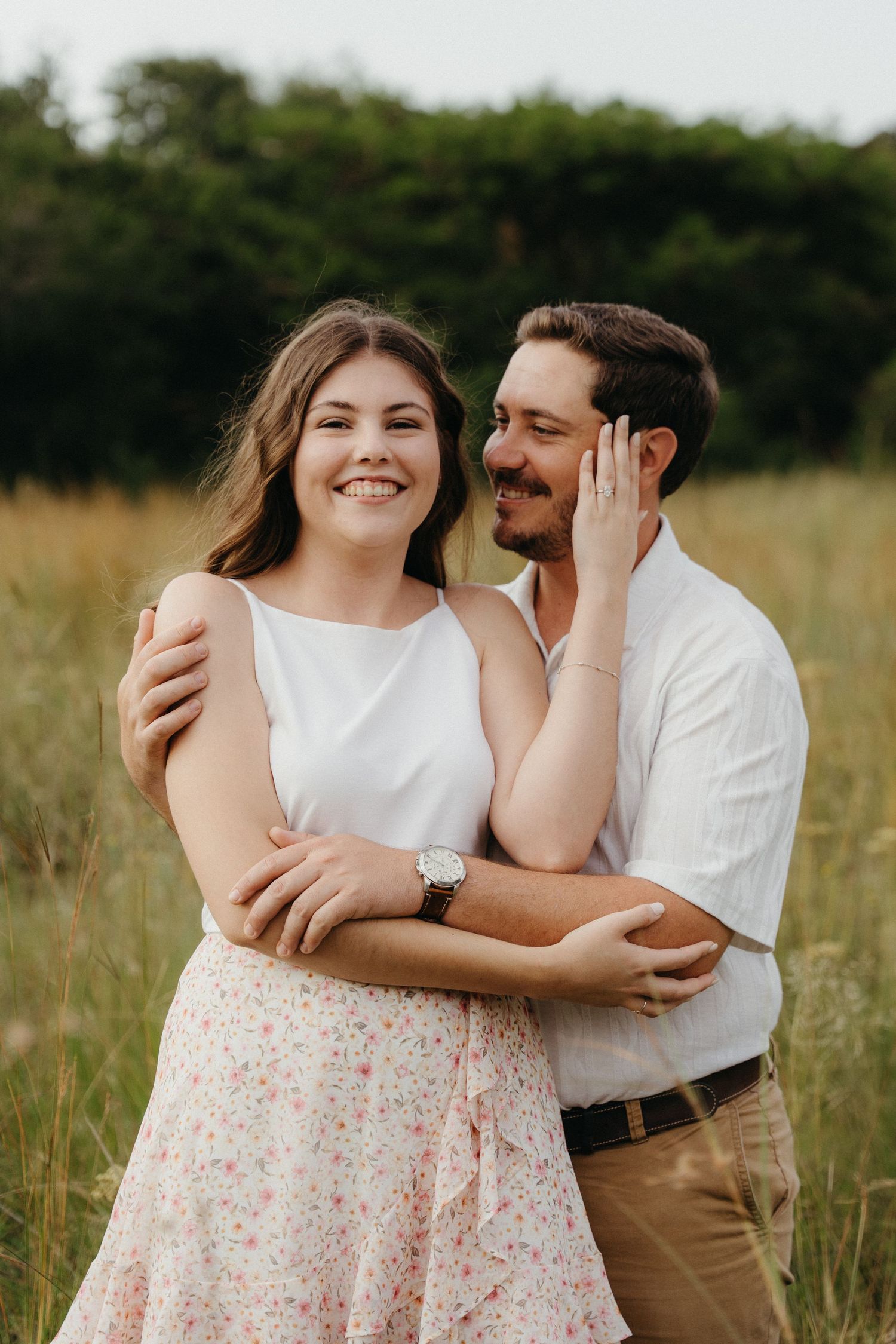 Couple embracing in a meadow, woman in white top and floral skirt smiling, man whispering in her ear.