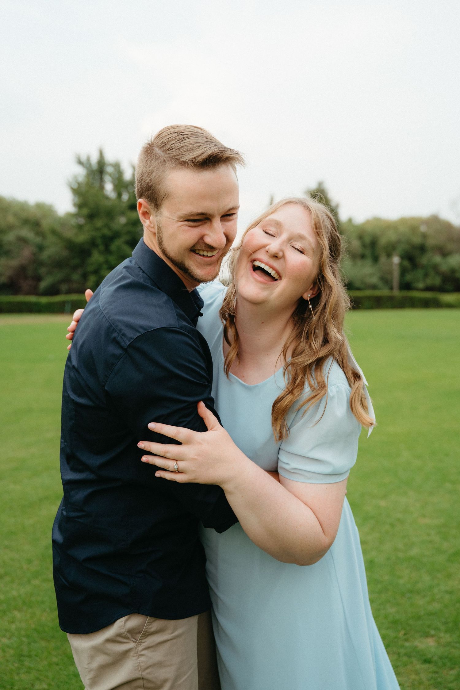 A couple shares joyful laughter while embracing on a green lawn during a sunny outdoor photo session.