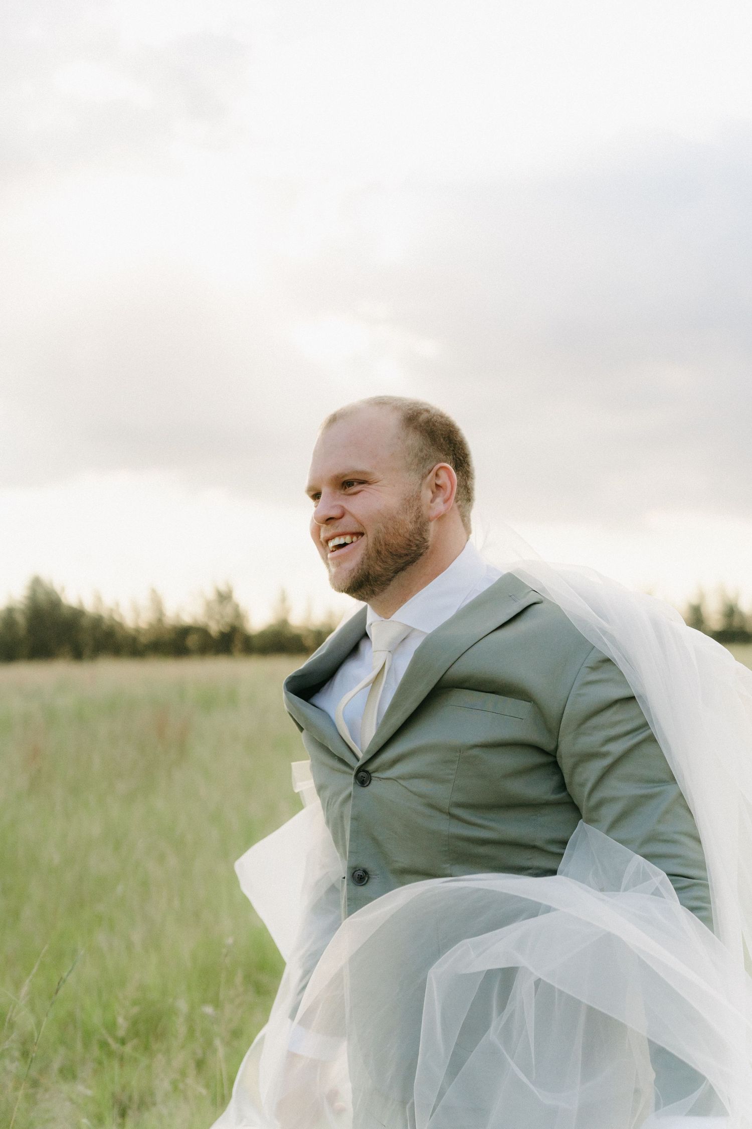 A groom wearing a light gray suit stands in a sunlit meadow with ethereal tulle fabric flowing around him.