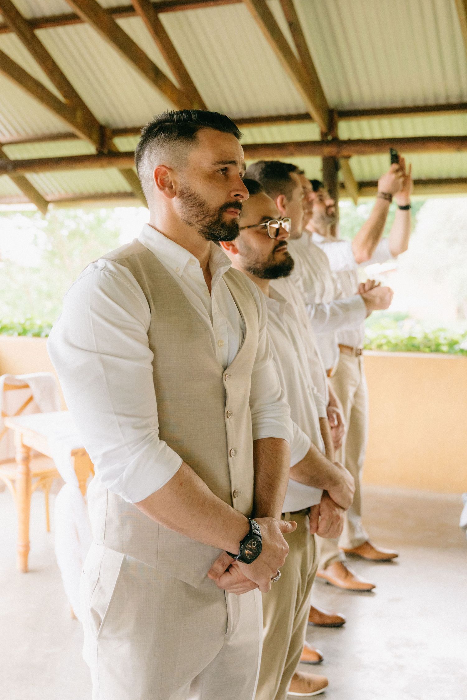 Groomsmen in light colored suits and white shirts gather under a covered outdoor venue.
