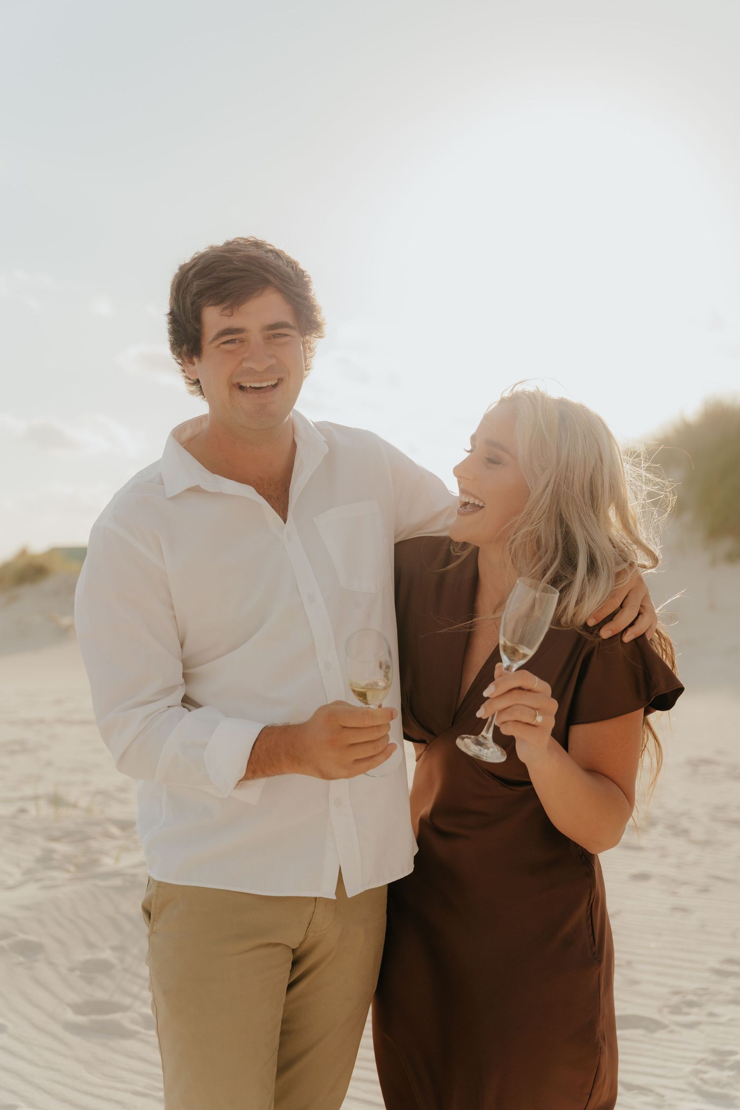 Couple laughing and holding champagne glasses on a sandy beach, man in white shirt and woman in brown dress.