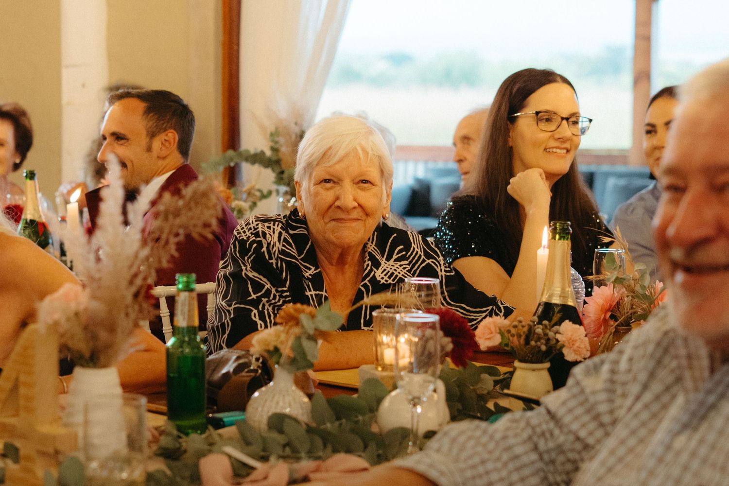 Wedding reception guests enjoying dinner and conversation at decorated table with warm lighting.