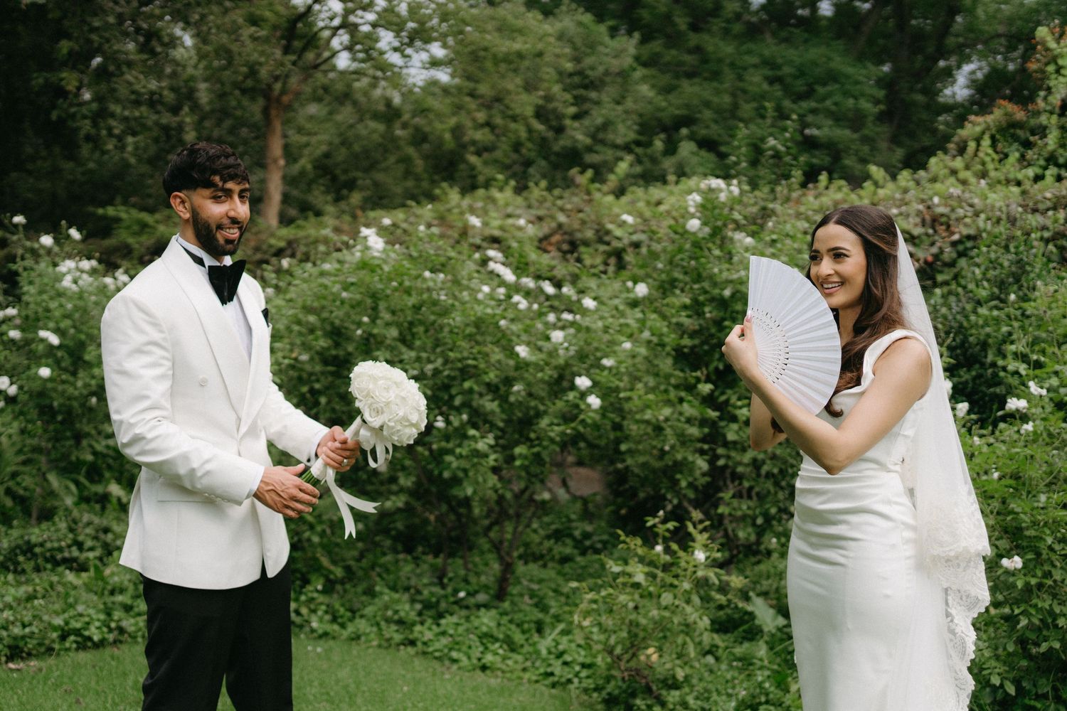 A couple in wedding attire sharing a playful moment in a garden with white flowers.