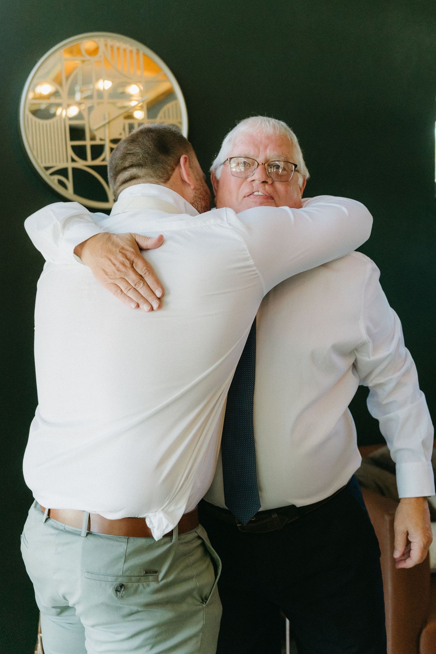 Two people in white dress shirts share an emotional embrace in front of a dark wall with a trophy display.