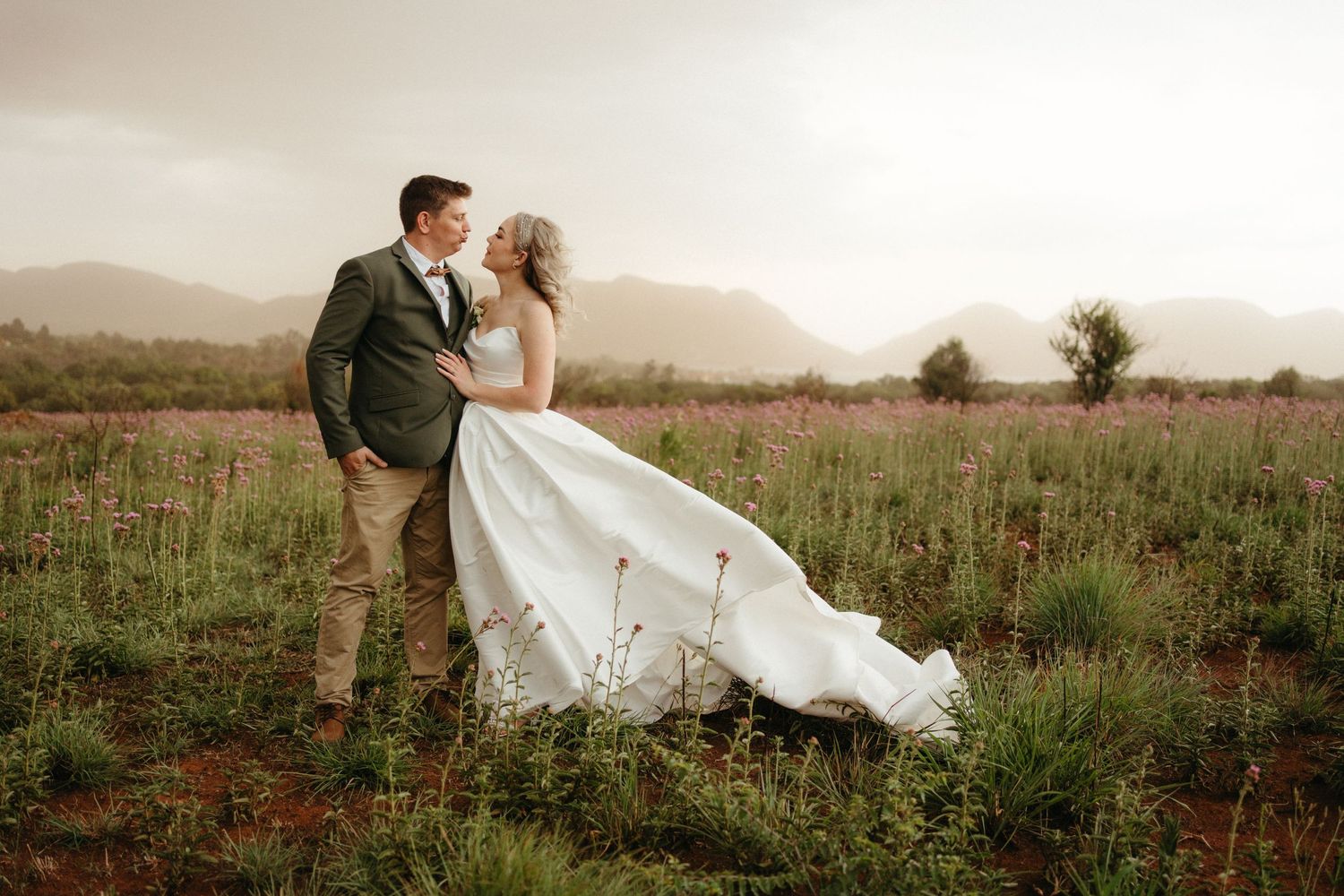 A couple in wedding attire embrace in a mountainside meadow as the bride's dress flows dramatically in the wind.
