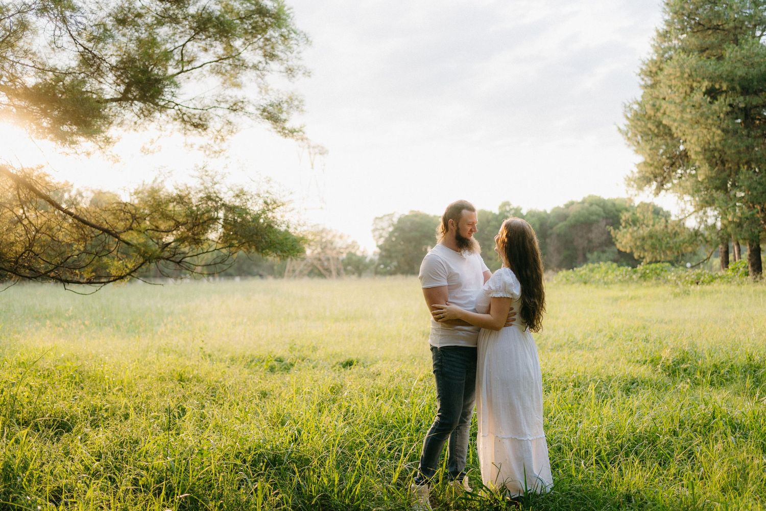 A loving couple embraces in a sunlit meadow with golden light filtering through trees at sunset.