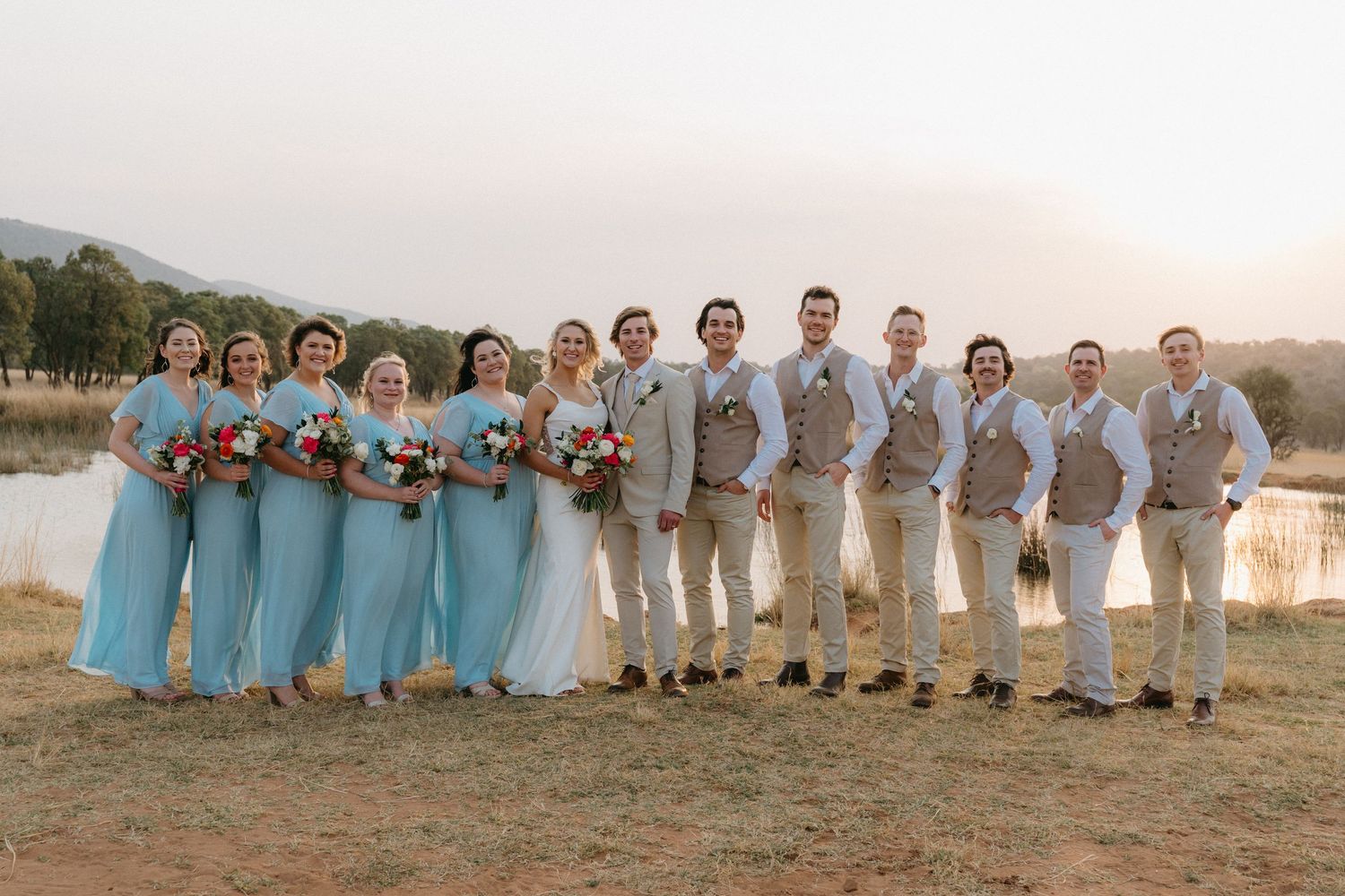 Wedding party in light blue dresses and tan suits pose together outdoors at sunset.