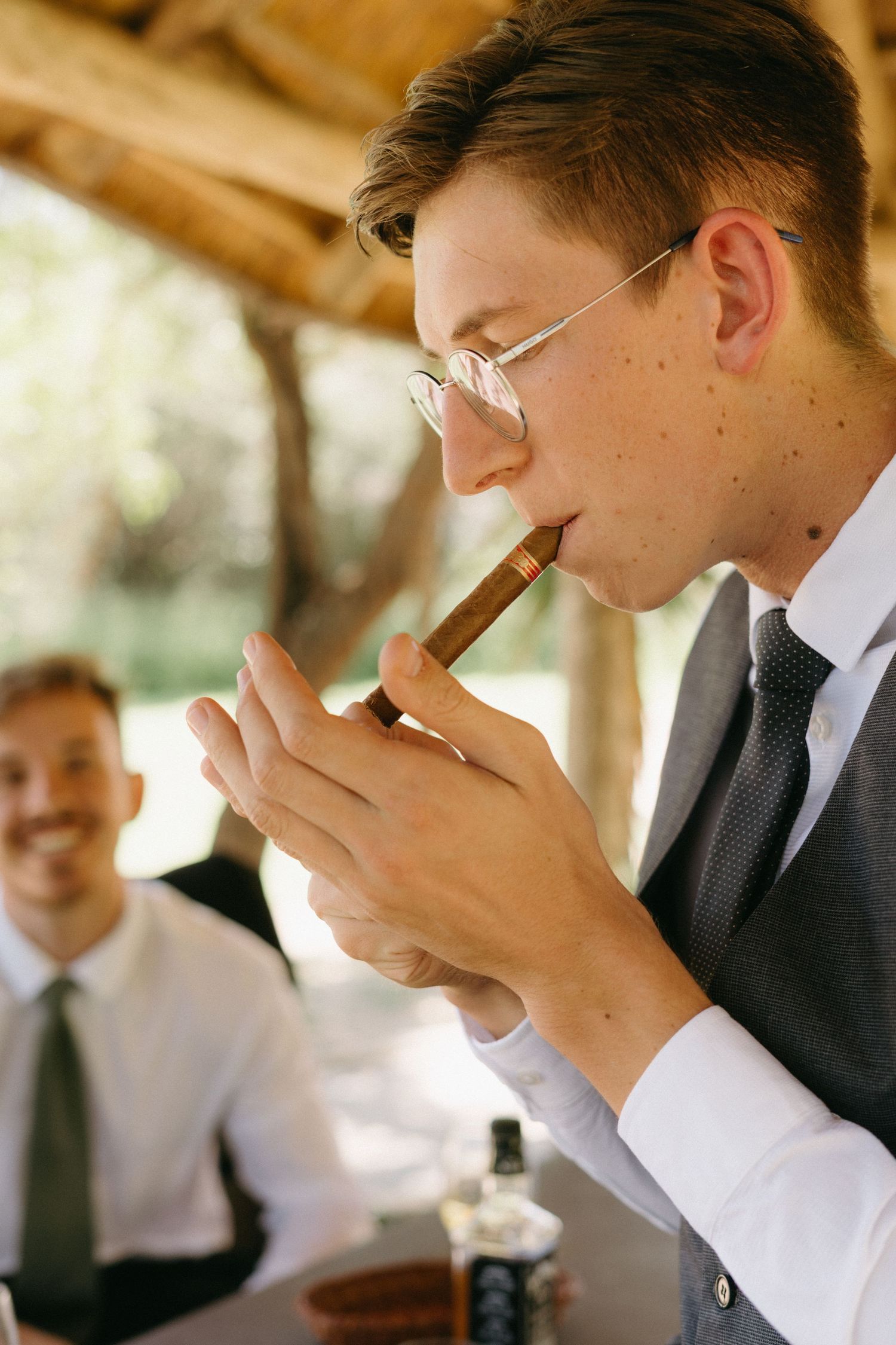Well-dressed man in suit and tie lighting a cigar during a celebration while another person smiles in background.