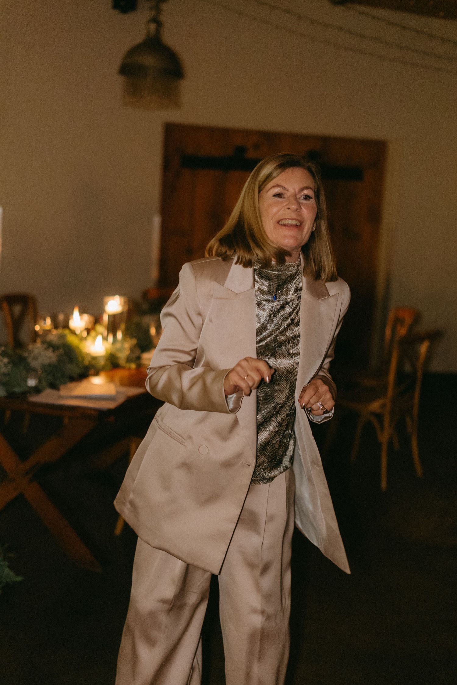 A person in a cream-colored suit dances joyfully at a wedding reception with string lights overhead.