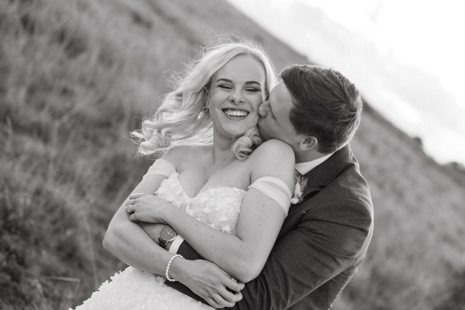 Black and white photo of a smiling bride being kissed on the cheek by groom in a field.