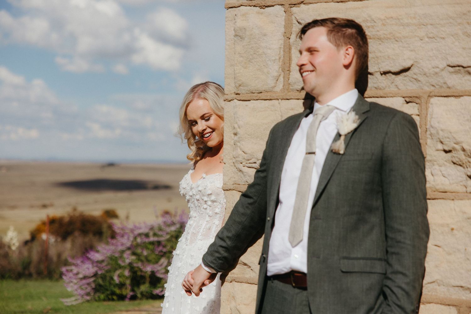 Bride and groom holding hands outdoors near stone wall, wearing wedding dress and green suit, with scenic countryside backdrop.