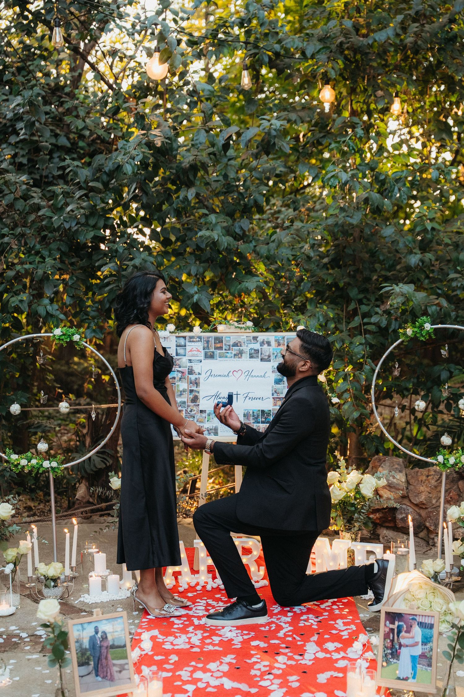 Man proposing on one knee to woman in black dress, surrounded by candles and floral decor in romantic garden setting.