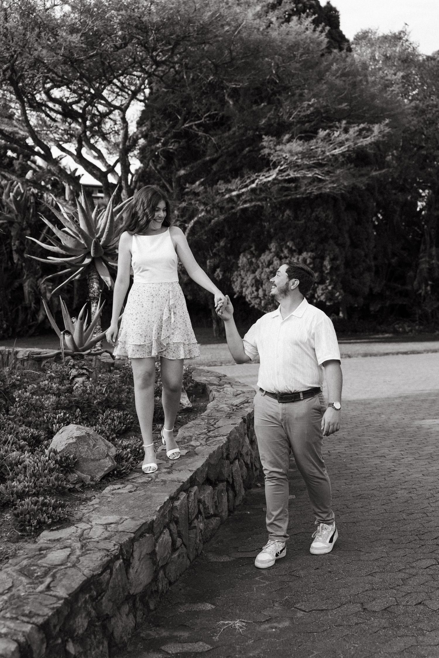 Black and white photo of a couple holding hands, walking along a stone wall path surrounded by lush trees and plants.