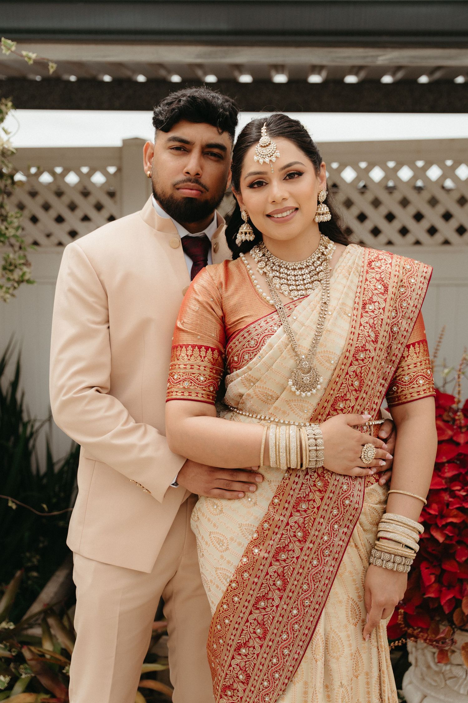 A couple in traditional South Asian wedding attire poses together, with the bride in a cream and red saree and groom in beige suit.