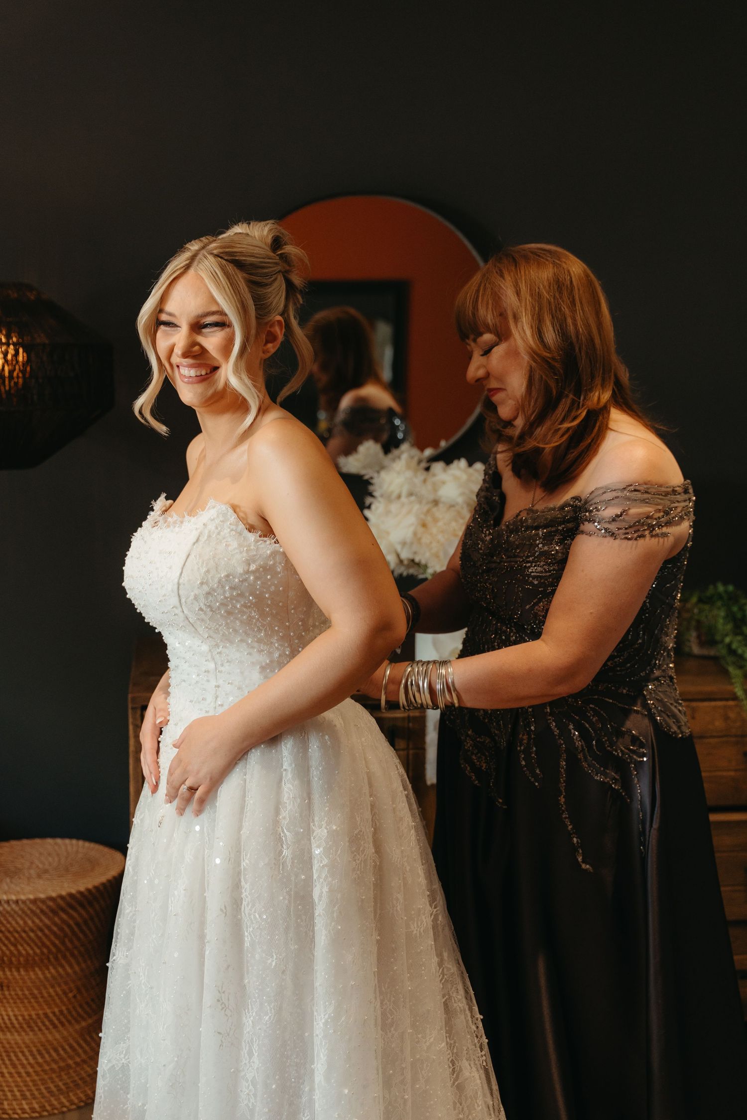 A bride in a white beaded wedding dress being helped with final adjustments before the ceremony.