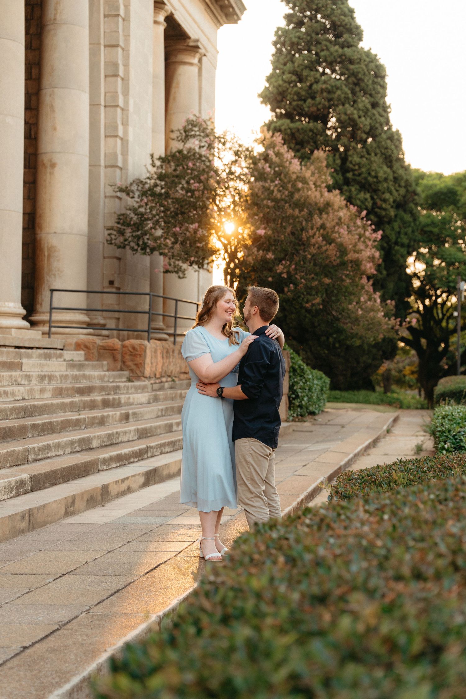 A romantic couple embraces on stone steps at sunset with classical architecture and cypress trees in the background.