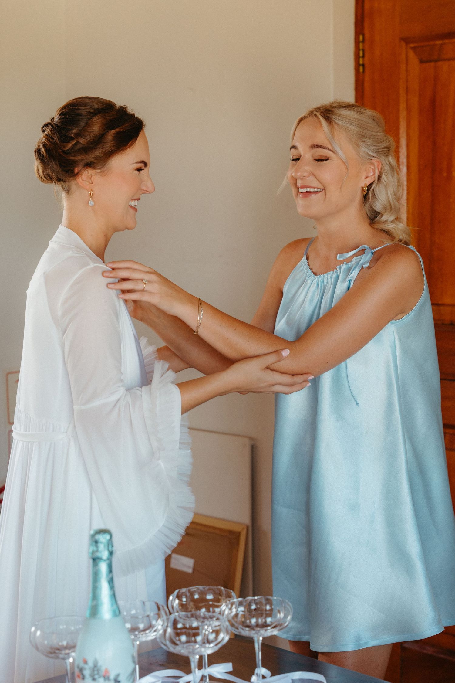 Two people in light blue and white outfits share a joyful embrace in an indoor setting with glassware on a table.