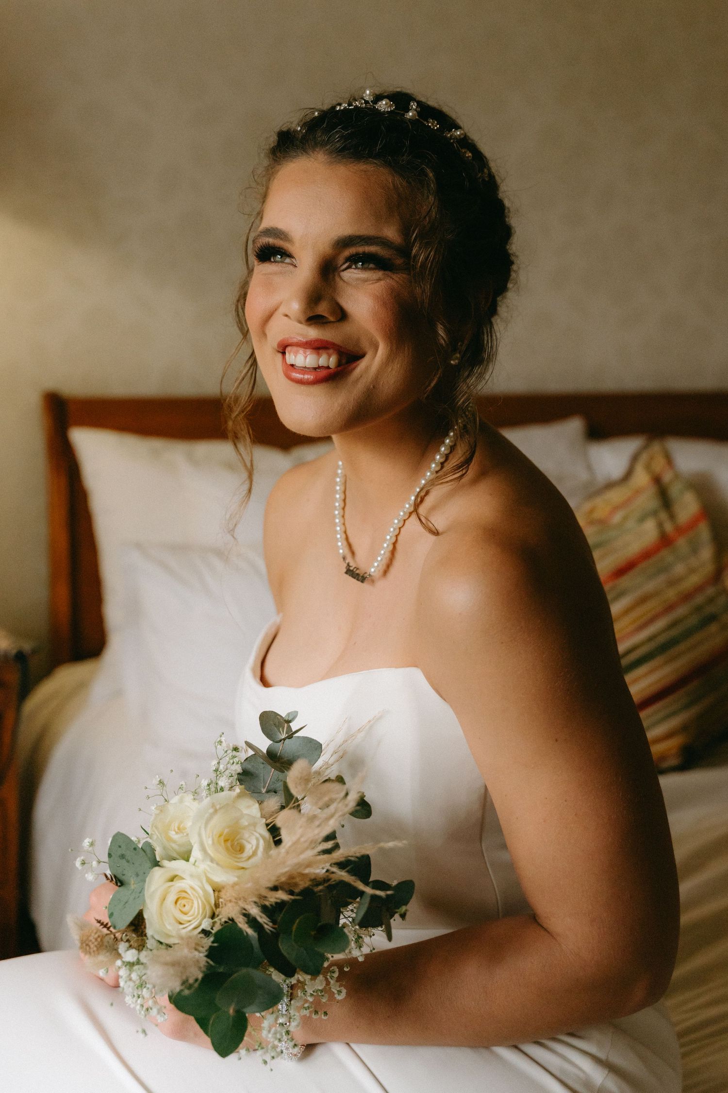 Bride in white dress holds elegant bouquet of white roses and eucalyptus while sitting indoors.