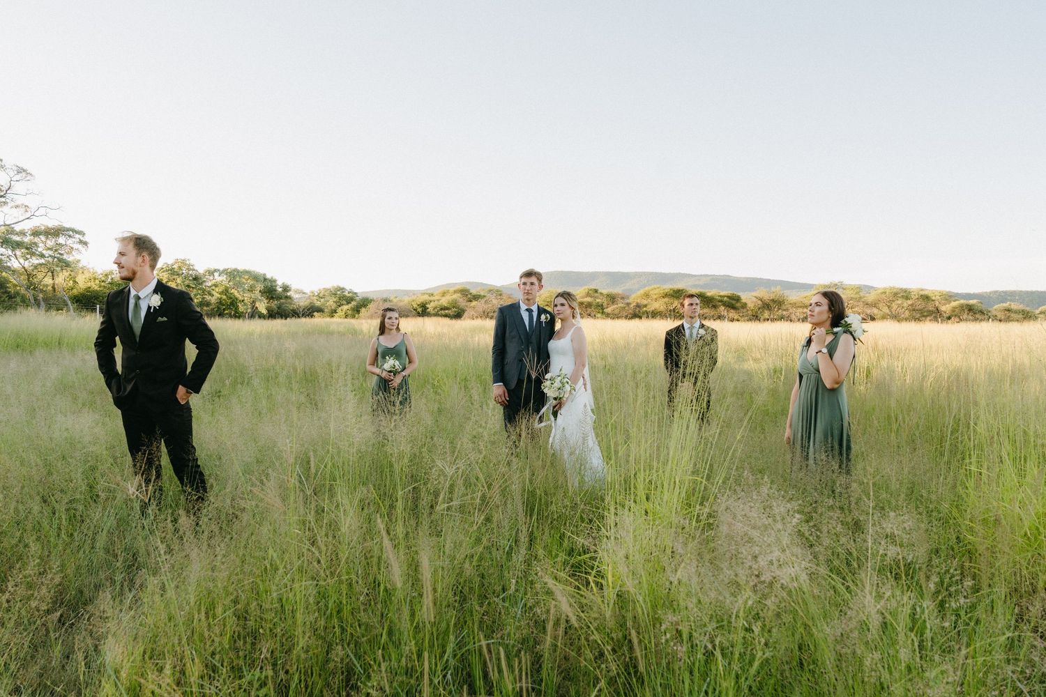 Wedding party standing spread out in a tall grass field under bright blue sky.