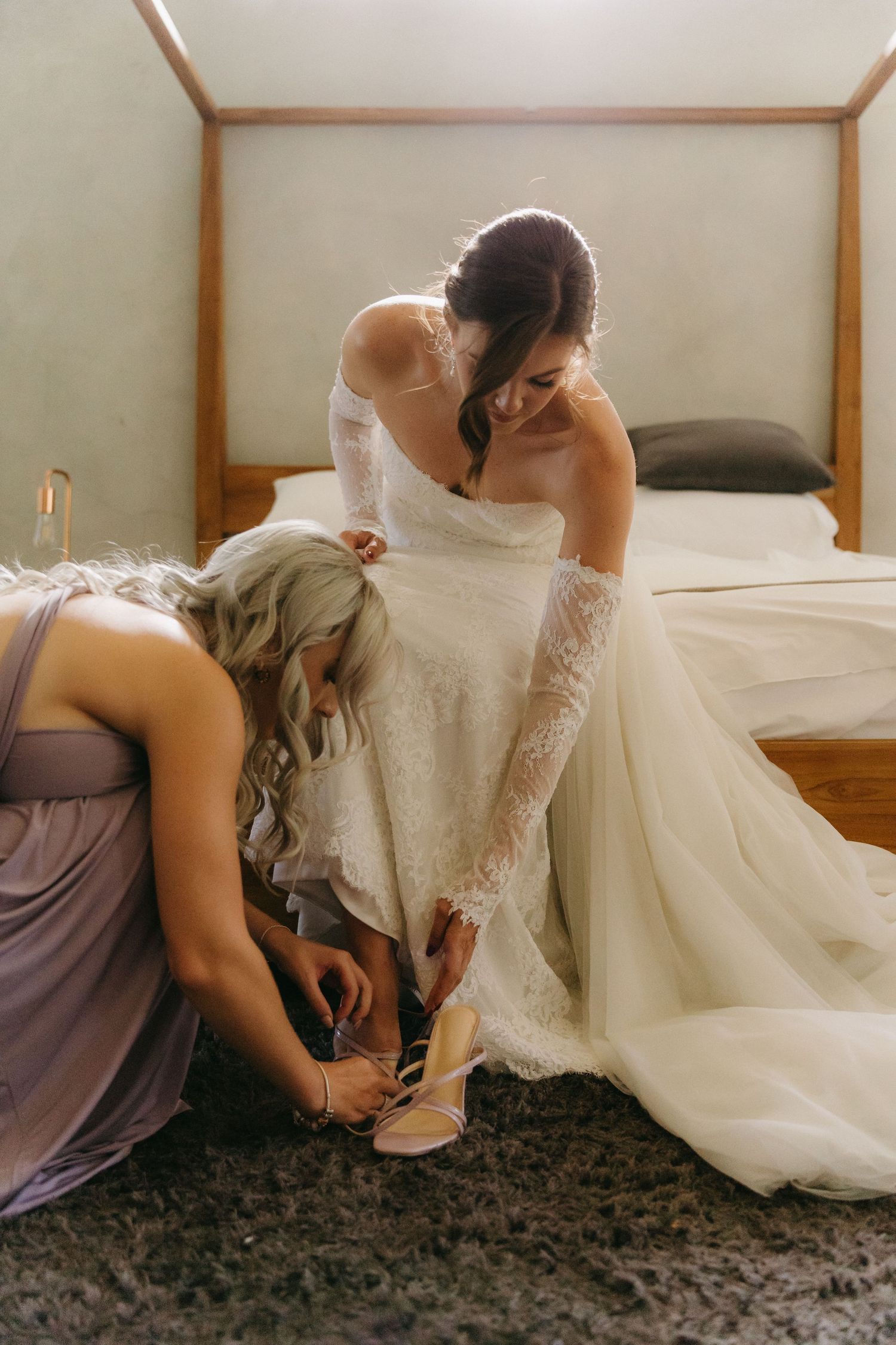 A bridesmaid in purple helps adjust a elegant lace wedding dress and shoes while getting ready on a wedding day.