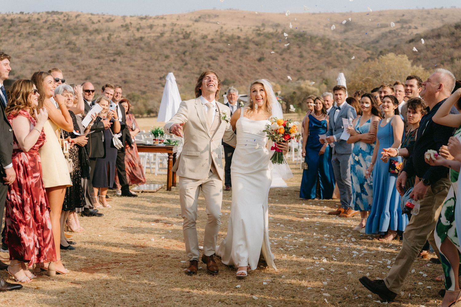 Guests gather outdoors at a desert wedding ceremony with rolling hills in the background.