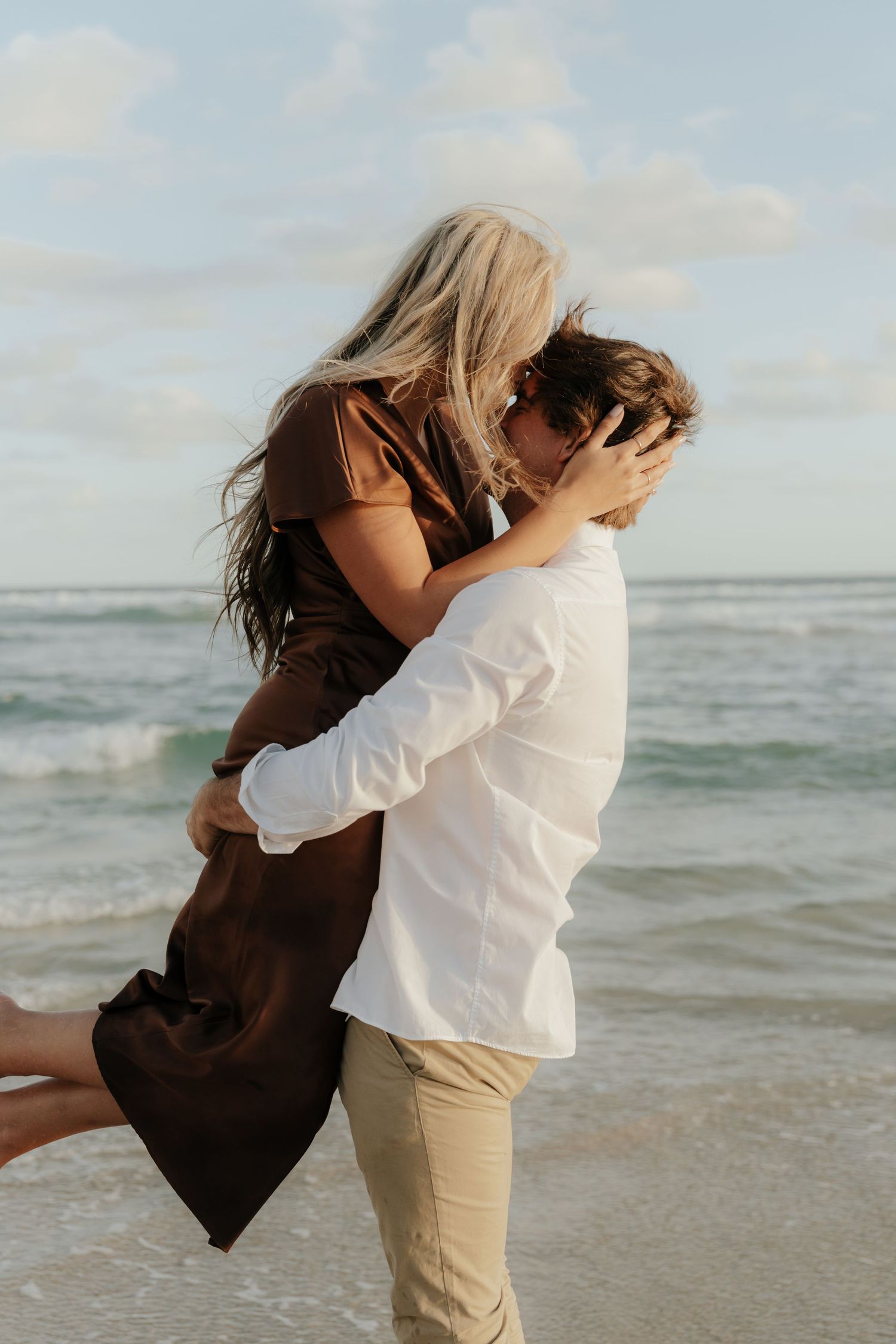 Couple embracing and kissing on a beach, woman in brown dress lifted by man in white shirt, ocean waves behind them.