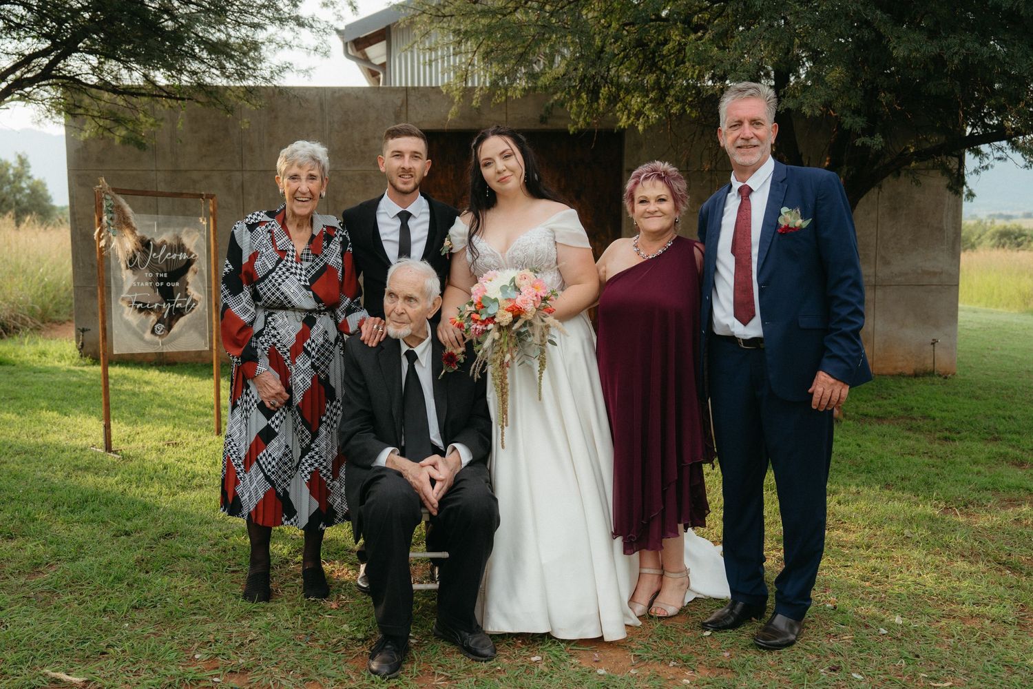 A wedding party poses together for a family portrait outdoors near a rustic building.