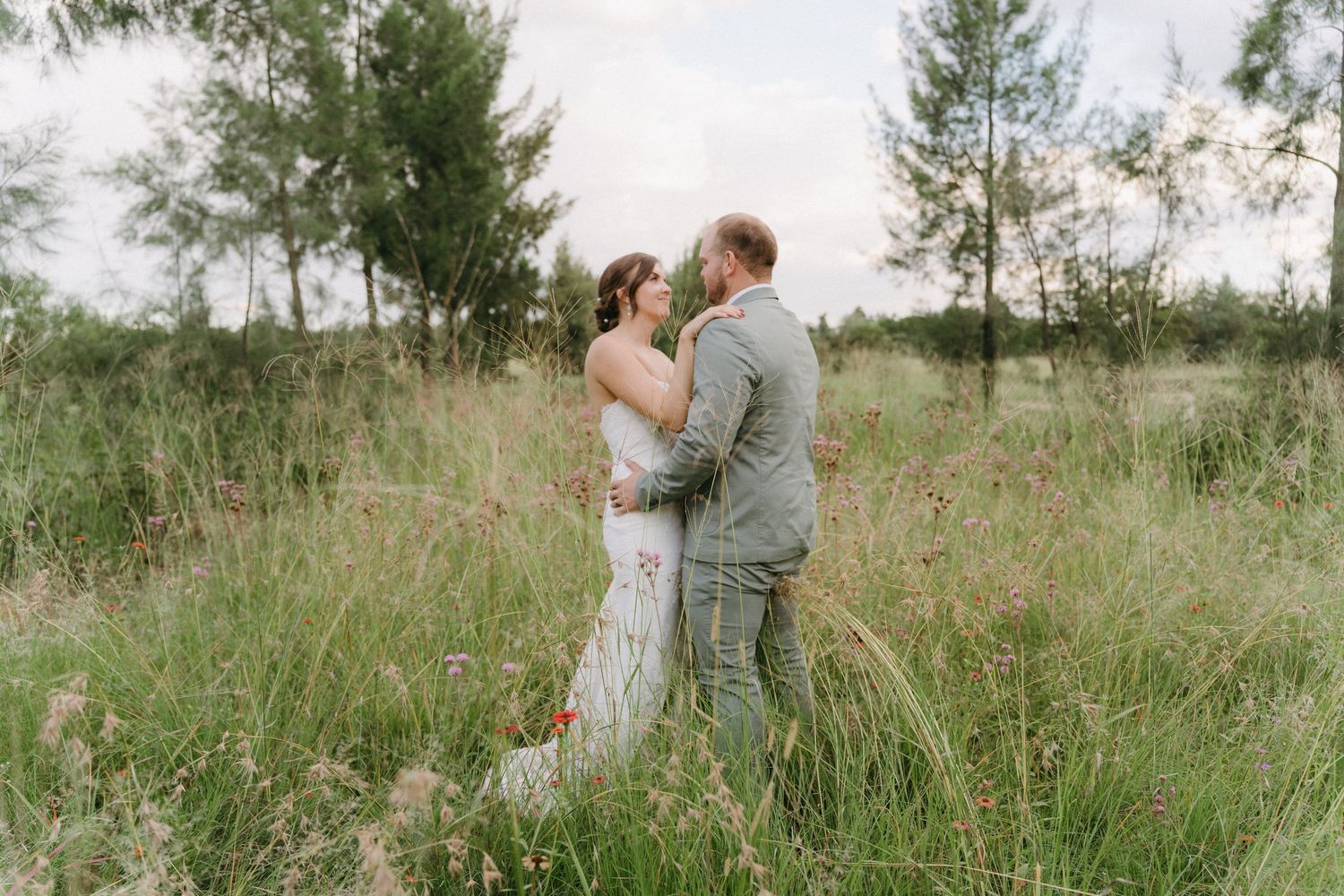 Newlyweds share an intimate moment while standing in tall grass with evergreen trees in the background.