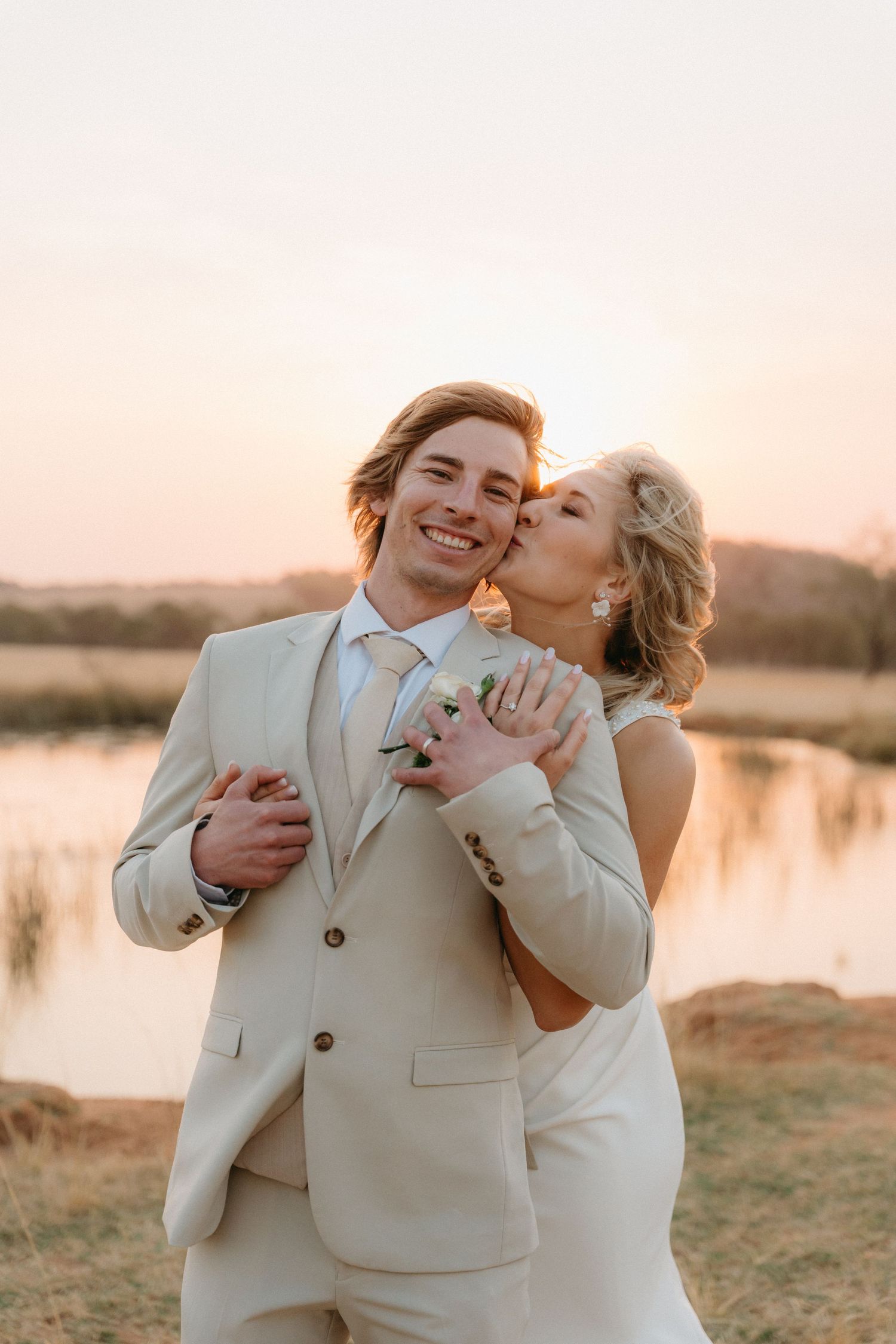 A romantic wedding couple embraces at sunset by a lake, wearing cream-colored attire in warm golden light.