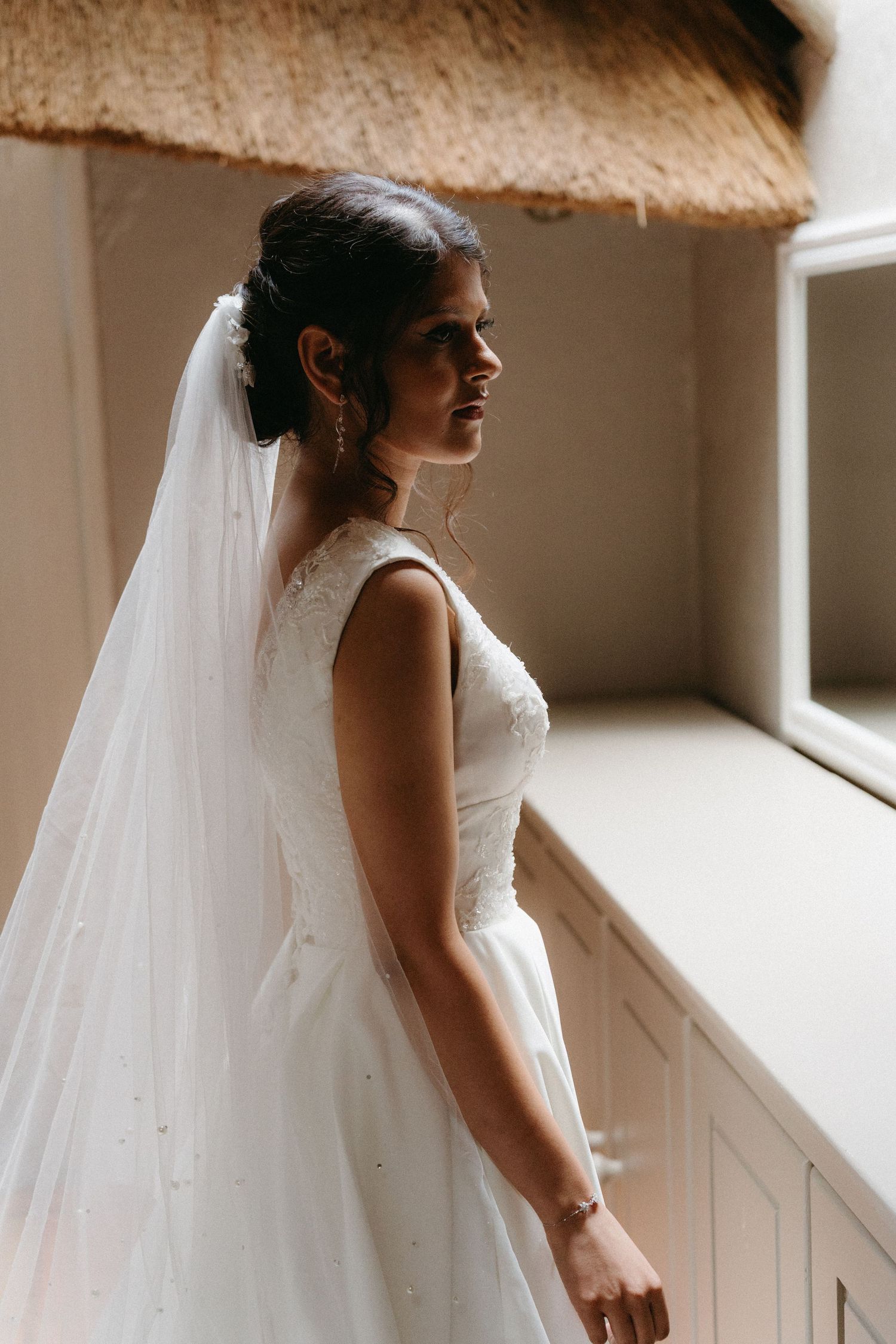A bride in a white sleeveless wedding gown and long veil stands in profile near a window in soft natural light.