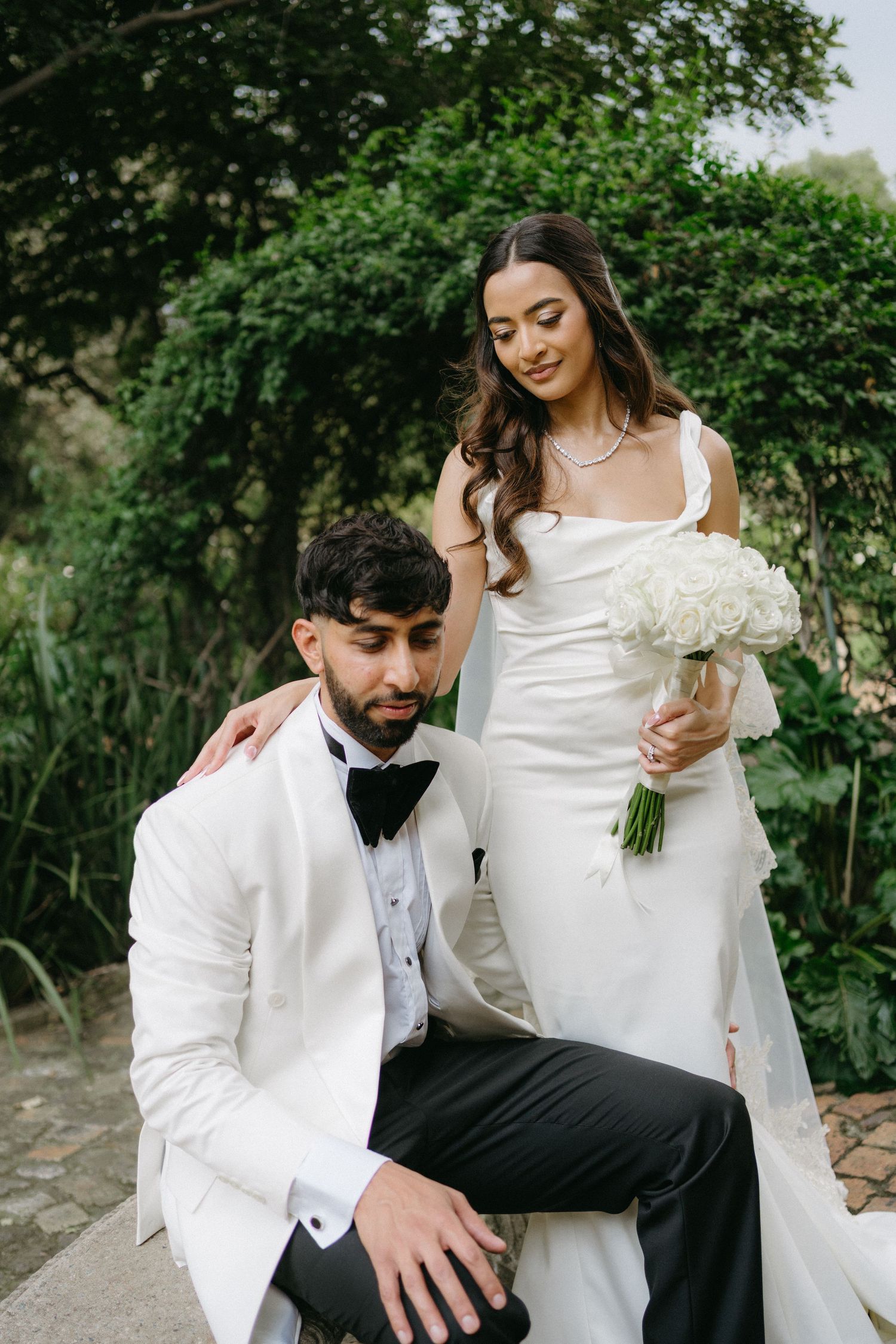 A wedding couple poses outdoors with the bride holding a white bouquet while the groom sits on a stone wall.