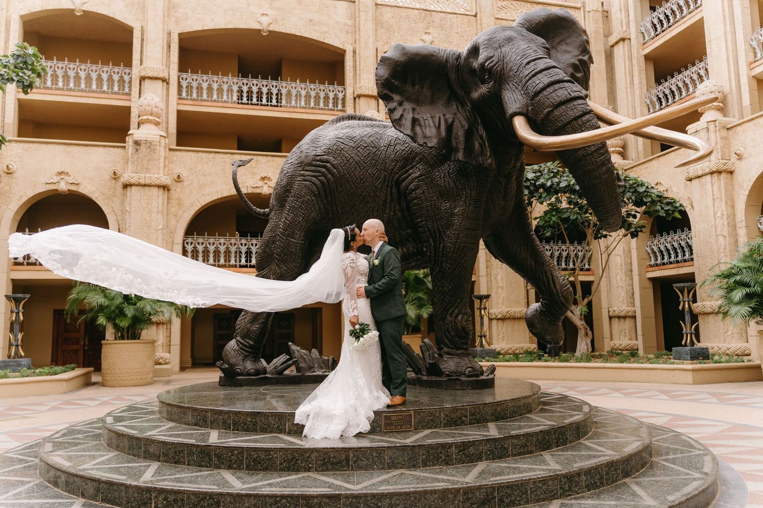 A dramatic wedding photo captured at a grand hotel courtyard featuring a large bronze elephant statue as centerpiece.