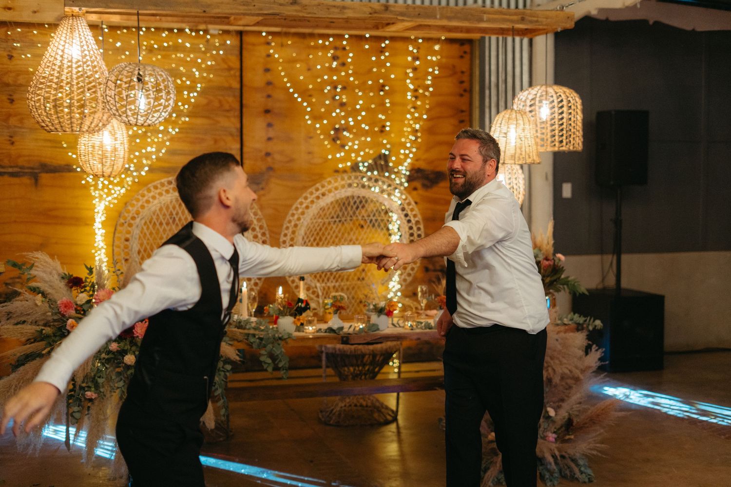 Two people dancing energetically at reception with fairy lights and rustic decor in background.