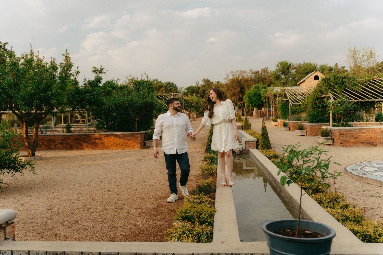 Happy couple holding hands walking through scenic garden pathway at Campagne Living with trees and water feature.