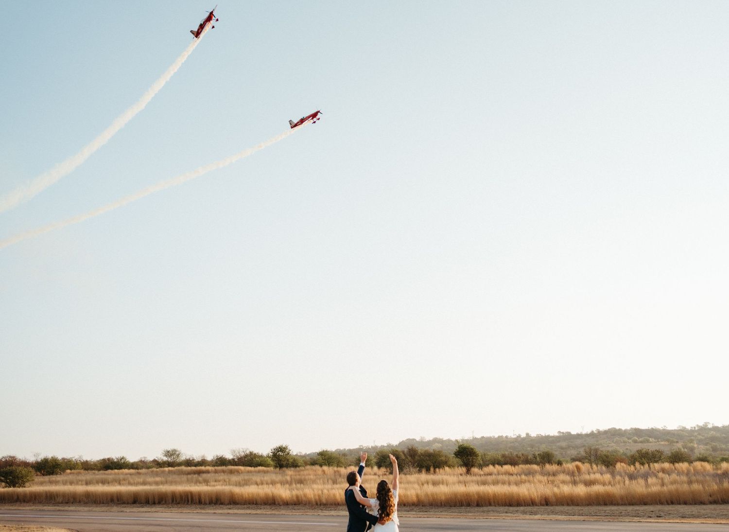 Red and white airplane trails streak across the sky above an open field during a wedding celebration.